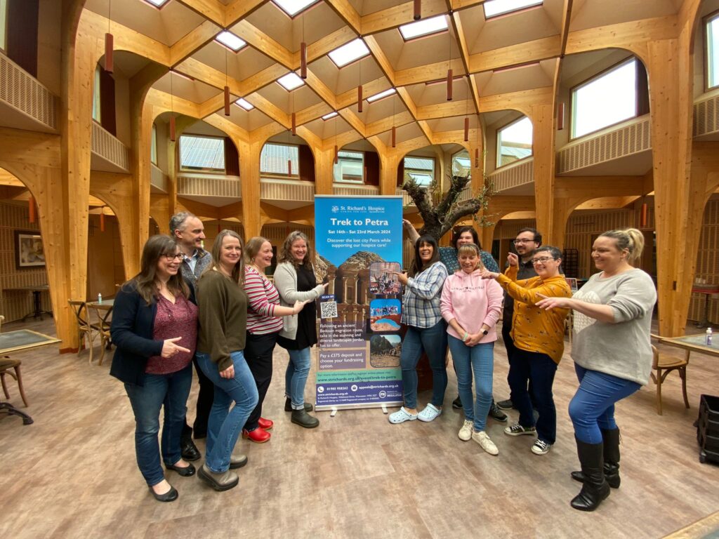 A group of people stand in The Green social space at St Richard's Hospice pointing at a tall banner featuring the Trek to Petra challenge wording and images.