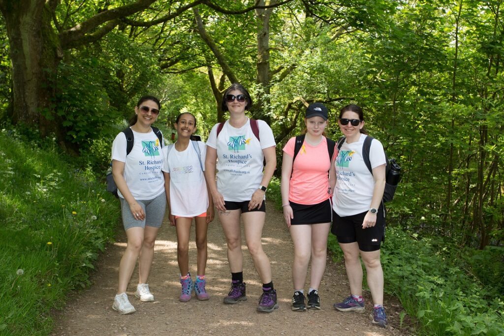 A group of people wearing St Richard's Hospice branded t-shirts stand together during the hospice's Malvern Hills Walk. They are on a pathway through a wooded area with a green canopy of trees.