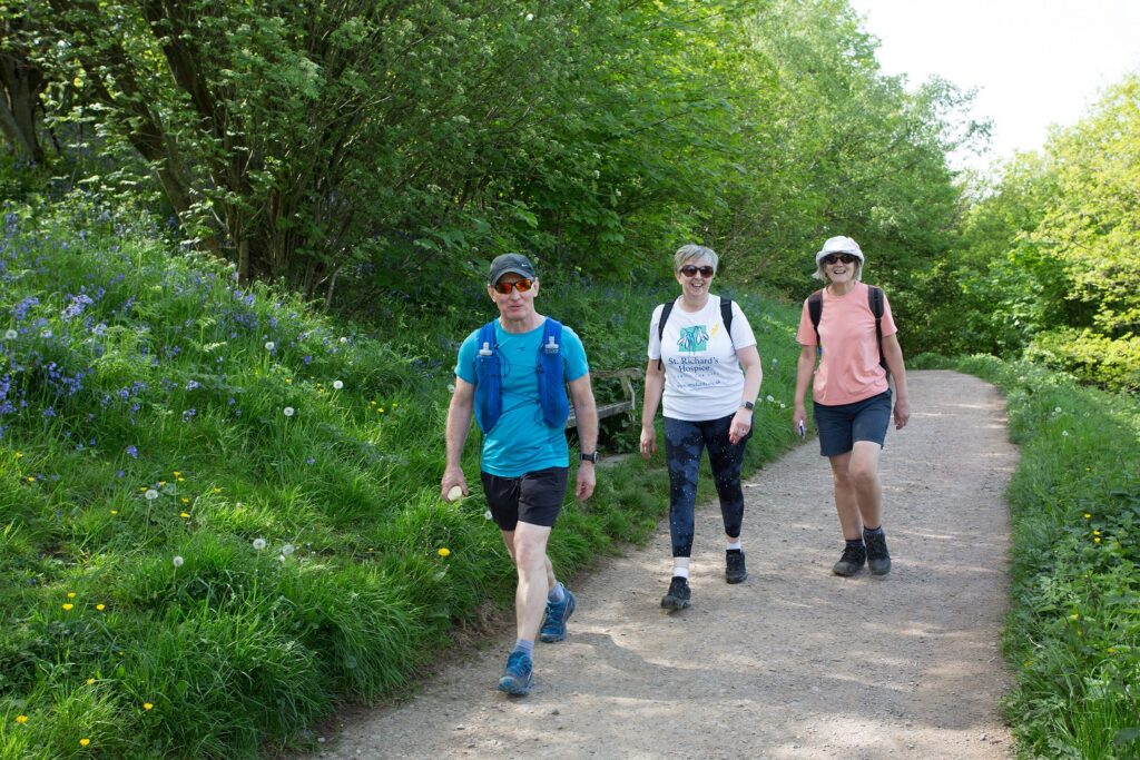 Three people in walking clothing stride along a path during the hospice's Malvern Hills Walk challenge. They are surrounded by vibrant greenery.