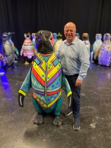 A man wearing jeans and a blue shirt stands smiling next to a penguin sculpture decorated to look like it's wearing an eighties tracksuit.