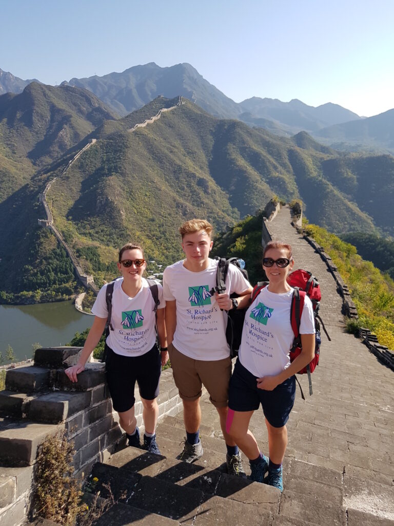 Three people wearing shorts and St Richard's branded t-shirts stand on the Great Wall of China. The wall stretches out behind them, along with rolling hills and blue skies.