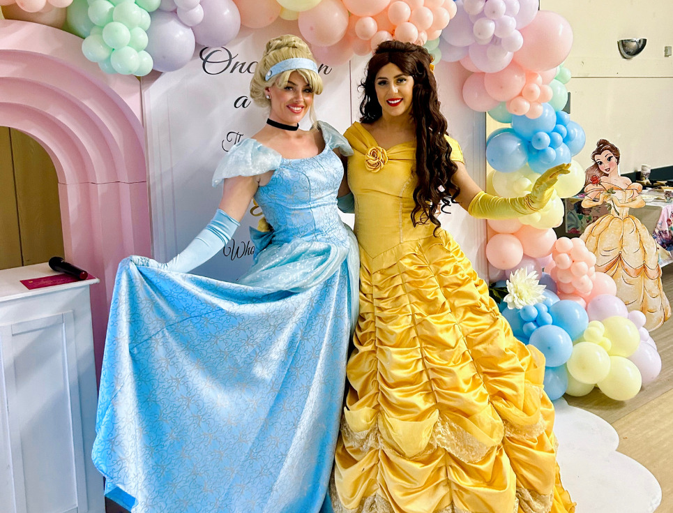Two people dressed as princess characters stand together in front of a pastel coloured balloon arch. They are wearing long, satin dresses. One dress is pale blue, the other is yellow.