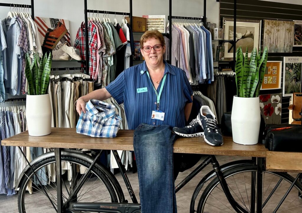 Sue stands in the hospice's store behind a display stand with bicycle wheels underneath holding up a folded shirt. On the display are two potted plants, a pair of jeans, and a pair of trainers.