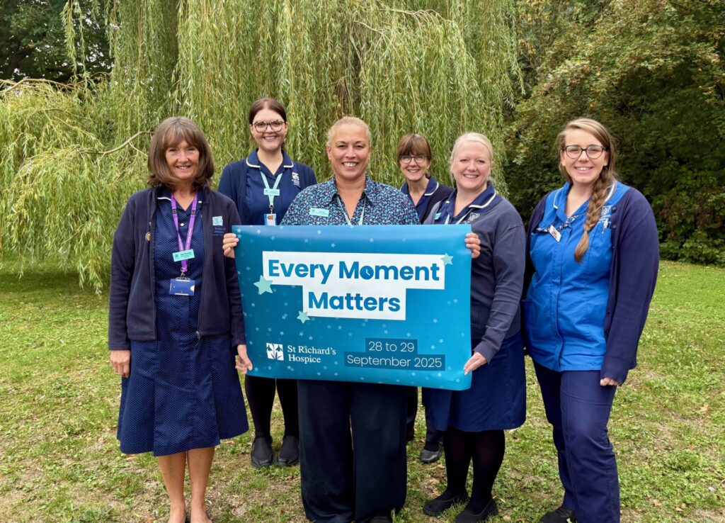 A group of nurses stand together in the hospice gardens holding a banner which reads Every Moment Matters.