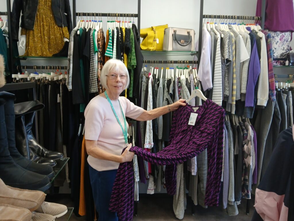 Volunteer Stella stands holding a dress she is about to put on a rail of clothes in the hospice's superstore. She is smiling.