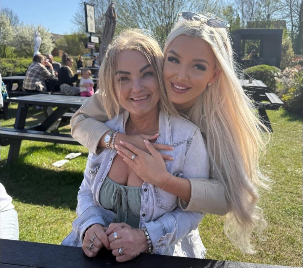 An older woman is seated at a picnic table in a sunny pub garden. A young woman embraces her from behind. They are both smiling.