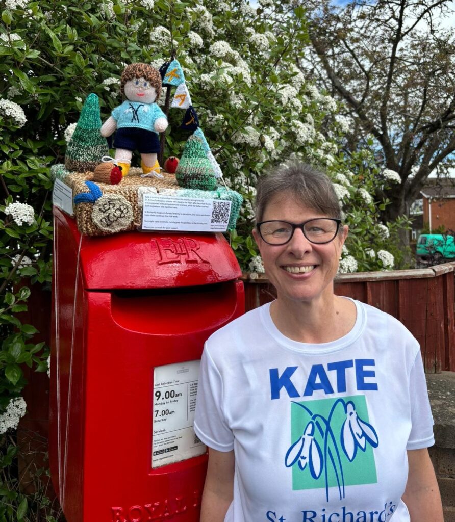 A woman standing beside a bright red Royal Mail post box decorated on top with a knitted figure, shrubs, and robins. The post box also displays an information card with a QR code. The person is wearing glasses and a white T‑shirt printed with blue and green text and graphics reading “KATE” and “St Richards.” Behind them are green shrubs with white flowers and a residential fence and houses.