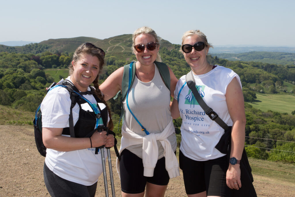 Three people wearing St Richard’s Hospice T-shirts stand together on a hilltop path, with countryside and rolling hills in the background.
