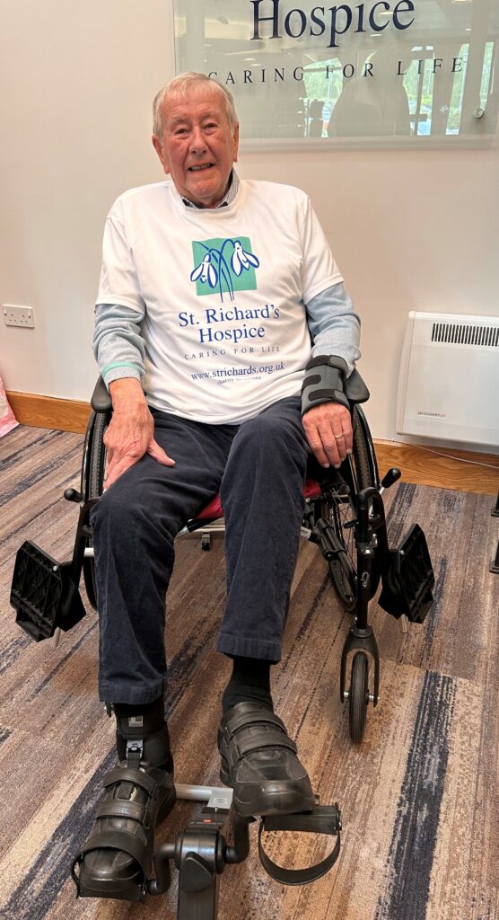 Person sitting in a wheelchair indoors, using a small cycle exerciser, wearing a St Richard’s Hospice T-shirt, with a hospice sign visible in the background.