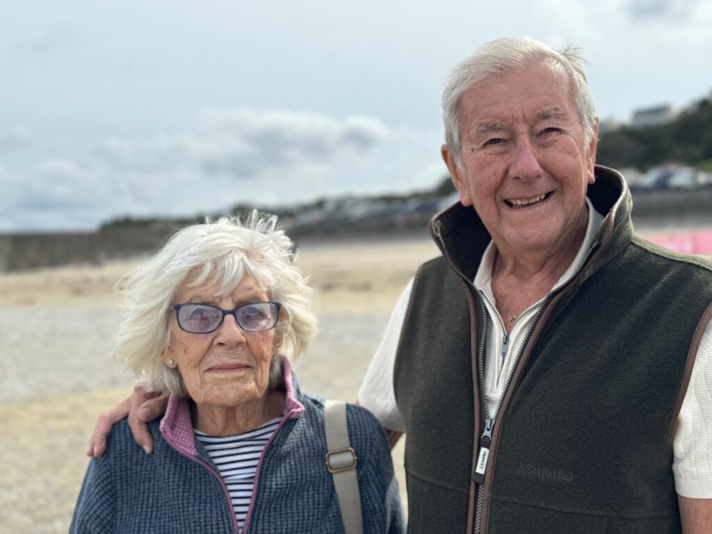 Older couple standing side by side on a beach, with one person’s arm around the other, and coastal buildings softly visible in the background.