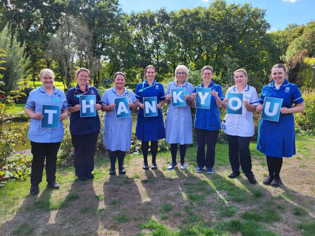 Eight members of the St Richard's Hospice team in uniform hold a thank you banner in the hospice gardens