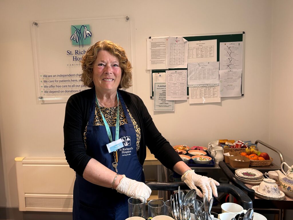 Volunteer preparing refreshments in the hospice kitchen, standing by a trolley with drinks, fruit and crockery at St Richard’s Hospice.