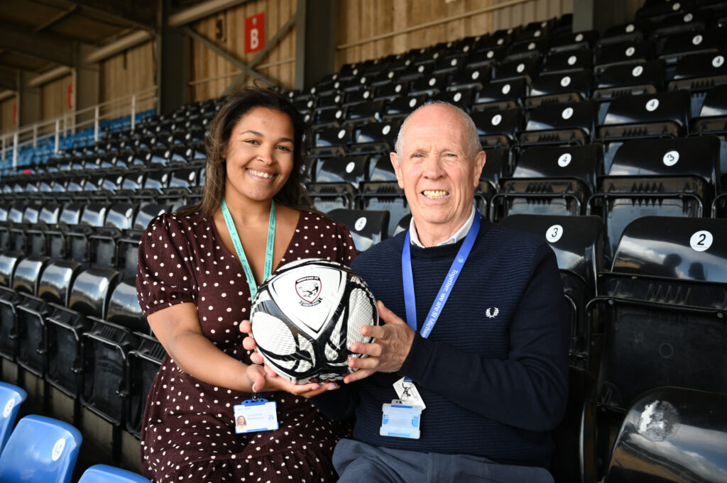 Two people sit in the stands of a football stadium holding a football between them. Both are wearing staff lanyards and are seated in front of rows of black stadium seats. The football displays a club crest, and the image appears to have been taken inside a covered spectator stand.