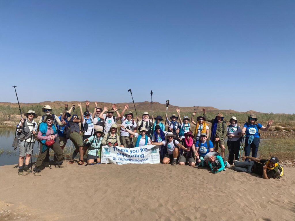 Fundraisers for St Richard’s Hospice pose with a thank you banner beside a river in a desert landscape after completing a trekking challenge.