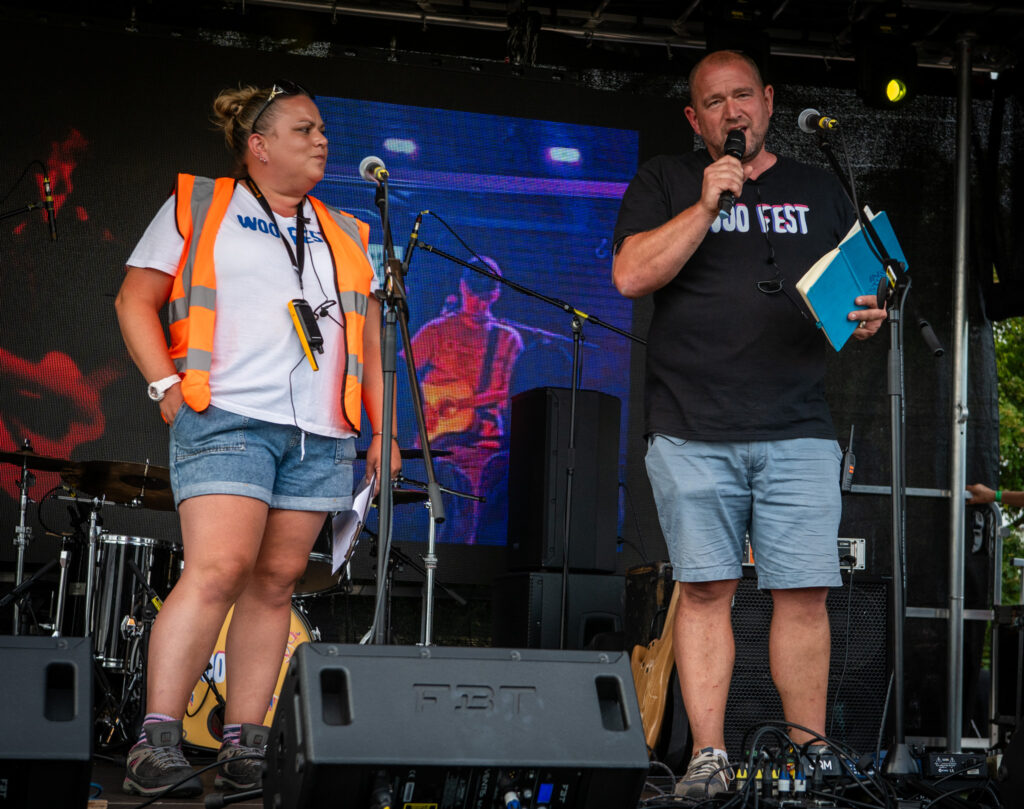 Two Woo Fest organisers speak into microphones on stage during the festival, with musical equipment and a large screen in the background.