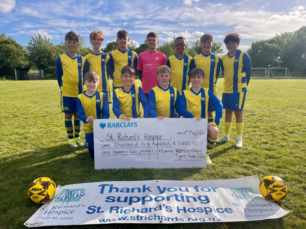 A youth football team in yellow and blue kits stands on a football pitch holding a presentation cheque for St Richard’s Hospice. The cheque shows a donation from Warndon Villages Tigers Under 12s. A St Richard’s Hospice banner and two footballs are displayed in front of the team, with goalposts and trees in the background under a bright blue sky.