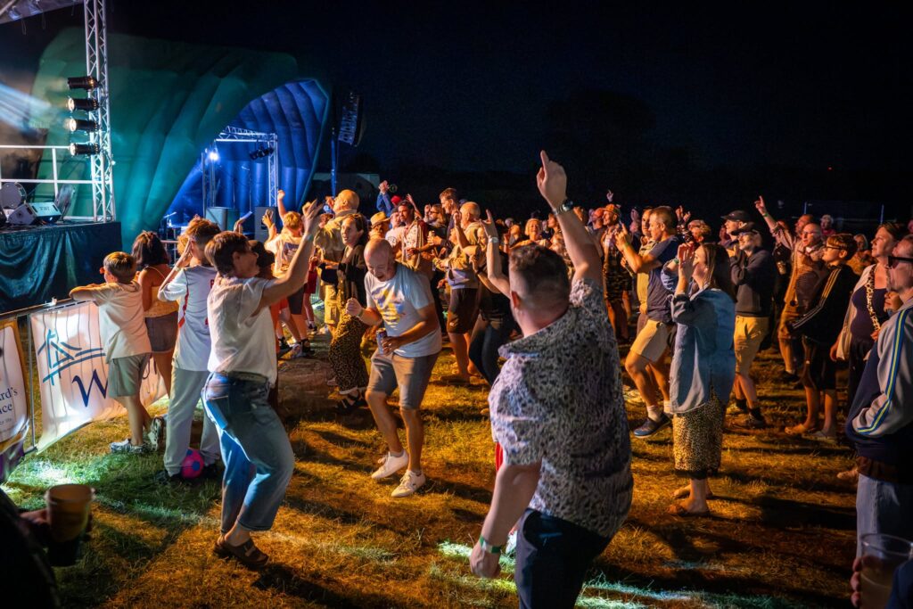 People dancing and enjoying live music in front of a brightly lit stage at an outdoor evening festival.
