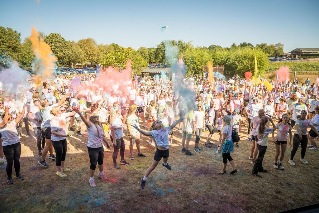 A large crowd of participants in white T-shirts throw brightly coloured powder into the air during a Rainbow Run event. Clouds of pink, blue, yellow and purple powder fill the outdoor space, with trees and blue sky in the background.