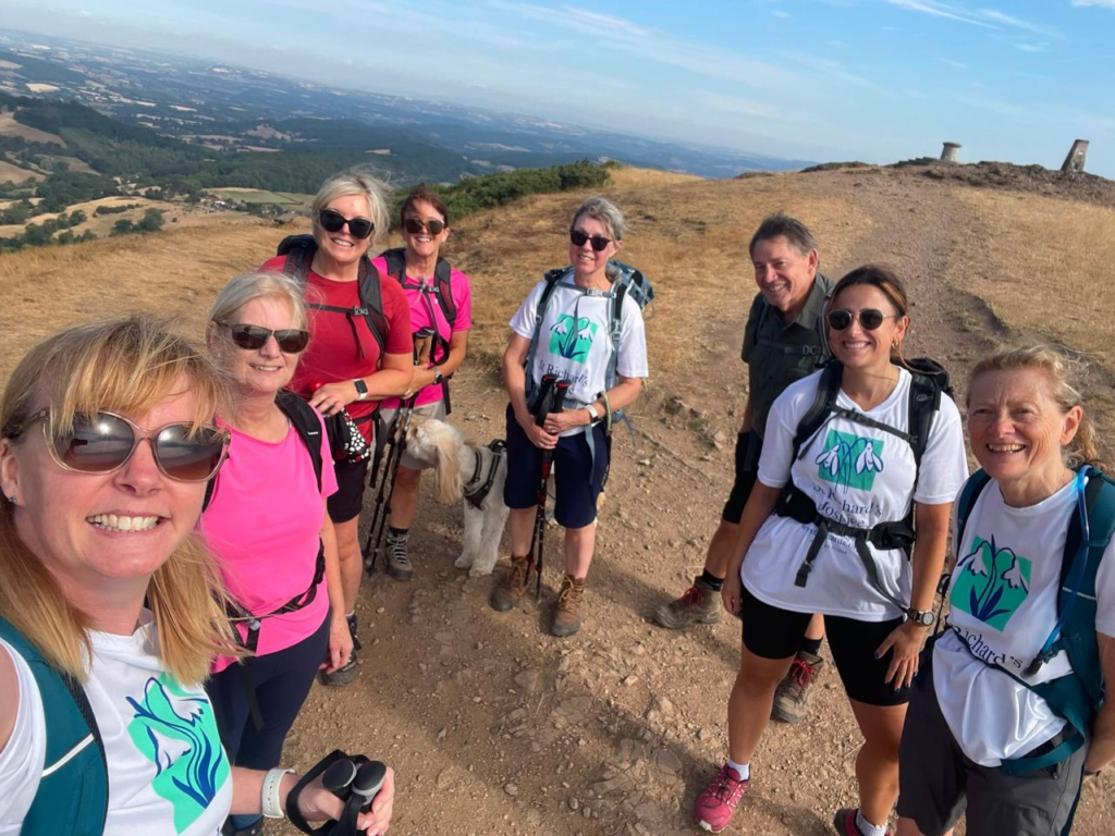 A group of eight charity supporters wearing walking gear and St Richard’s Hospice T-shirts pause for a photo on a training walk in the Malvern Hills. The group stands on a hillside path with wide views of the Worcestershire countryside in the background. They are training to walk the Great Wall of China.